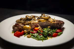 Juicy grilled steak topped with mushrooms, served with fresh vegetables on a white plate.