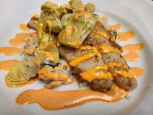 Grilled fish fillet topped with vegetables and drizzled with spicy sauce, served on a white plate.