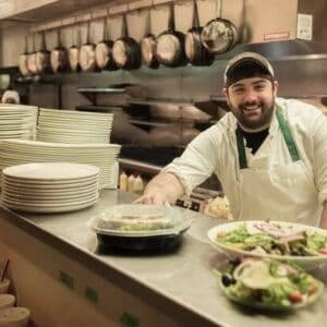 Happy chef preparing fresh salads in a busy restaurant kitchen.