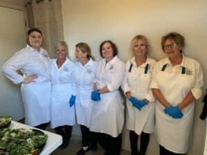 Group of women chefs in white uniforms and blue gloves preparing salads.